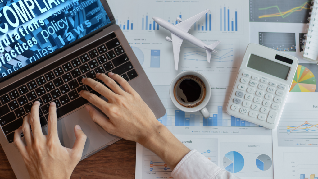 Image of a busy desktop with hands working on a laptop, business reports, coffee cup, calculator. Laptop screen displays words such as compliance, policy, regulations, and laws. 